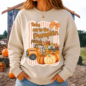 May include: A sand-coloured crewneck sweatshirt featuring a graphic print of an orange vintage truck loaded with pumpkins. The text reads "take me to the Pumpkin patch" in orange script, accompanied by a plaid bow and a striped rectangular frame.