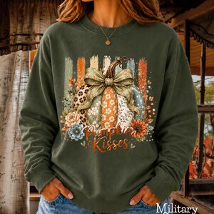 Peut inclure: Un sweat-shirt à col rond vert militaire orné d'un motif de citrouille avec un grand nœud, entouré d'éléments floraux et de coups de pinceau verticaux. Le texte "Pumpkin Kisses" est imprimé en écriture cursive orange sous la citrouille.
