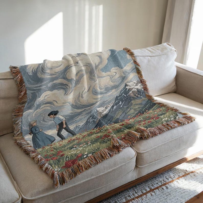 May include: Woven throw blanket featuring a mountain landscape with two figures. The blanket has a fringed edge and depicts a scene with a cloudy sky, snow-capped mountains, and a field of red flowers. The color palette includes blues, greens, and browns.