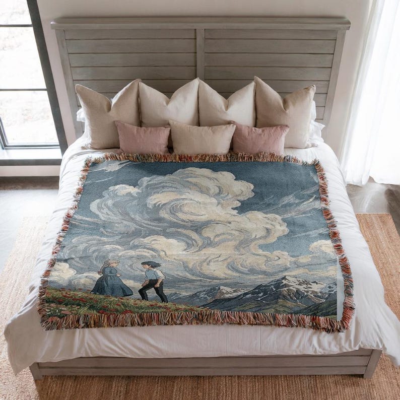 May include: A woven throw blanket featuring a landscape scene with a couple walking on a hill, under a cloudy sky. The blanket has a blue and white color palette with a fringed edge. The throw is placed on a bed with pillows.