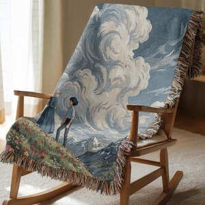May include: A woven throw blanket featuring a landscape scene with two figures, mountains, and clouds in shades of blue, gray, and beige. The blanket has a fringed edge and is draped over a wooden rocking chair.