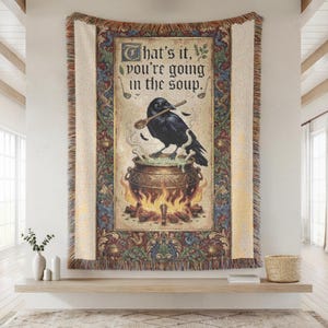 May include: A woven throw blanket featuring a black raven perched on a cauldron over a fire, holding a wooden spoon. The text reads, "That's it, you're going in the soup." The design is framed by an ornate, colorful floral border with fringe.