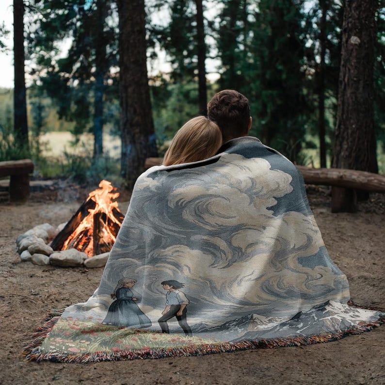 May include: A woven blanket with a cloud design in shades of gray and blue, draped over two people. The blanket features a scene with two animated characters and a mountain landscape. The blanket has a fringed edge and is set outdoors near a campfire.