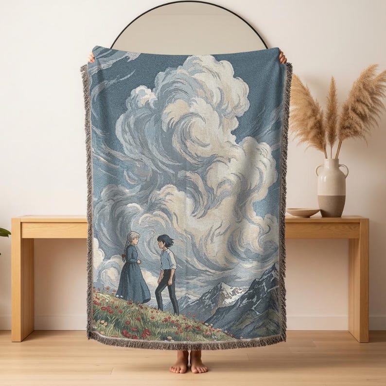 May include: A woven throw blanket featuring a scene with two animated characters standing on a hillside with mountains in the background. The blanket has a blue and white color scheme with a fringed edge.