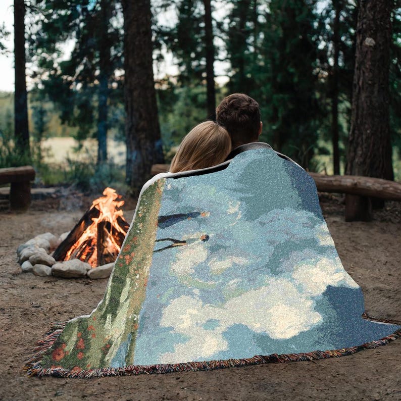 Puede incluir: Una manta tejida con un dise&ntilde;o de paisaje, sobre dos personas cerca de una fogata. La manta presenta un cielo azul con nubes, una ladera verde y un fleco. La escena est&aacute; ambientada al aire libre.