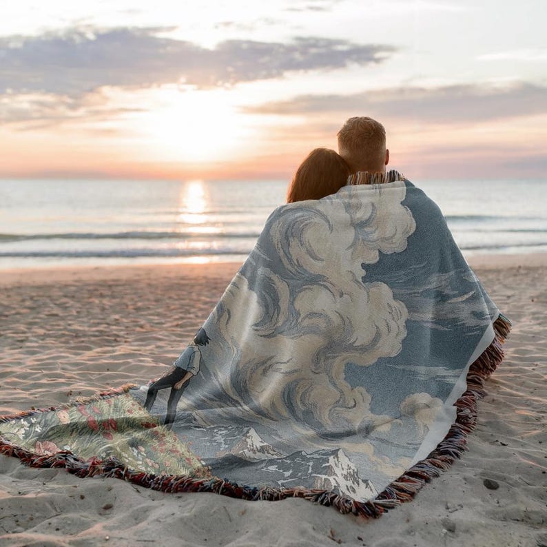 May include: A woven blanket with a landscape design featuring clouds, mountains, and a figure, draped over two people on a beach. The blanket has a blue and white color scheme with a fringed edge. The scene is set at sunset.