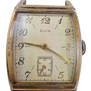 Op de afbeelding: Vintage Elgin polshorloge met een rechthoekige goudkleurige kast. De wijzerplaat is gebroken wit met zwarte cijfers en wijzers, inclusief een kleine secondenwijzer. Het woord "ELGIN" staat op de wijzerplaat.