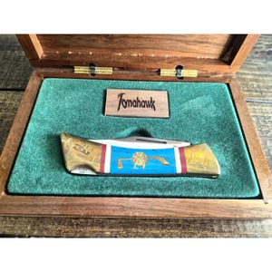 May include: A folding pocket knife with a gold-toned handle featuring a blue center section with a gold emblem, resting in a wooden display box with green velvet lining. A small wooden plaque inside the box displays the text Tomahawk.