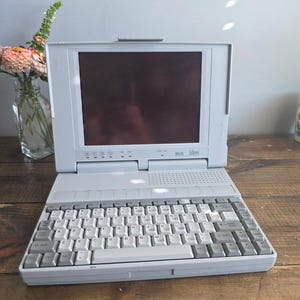 Puede incluir: Un ordenador portátil vintage Librex 286 SX de color gris claro con pantalla oscura, un teclado QWERTY completo con teclas de color gris claro y una serie de luces indicadoras de estado situadas bajo la pantalla en el chasis.