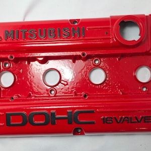 Może przedstawiać: Jasnoczerwona pokrywa zaworów silnika z logo Mitsubishi i czarnym napisem "DOHC 16 VALVE". Pokrywa posiada kilka okrągłych otworów i otwory montażowe.