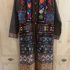 May include: A long-sleeved jacket featuring a dark brown base with intricate, multi-coloured embroidered panels. The front displays vibrant geometric patterns, small circular mirrors, and a central button closure. The sleeves have a dark brown patterned fabric.