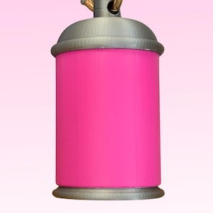 Op de afbeelding: Een felroze spuitbus sleutelhanger met een zilveren dop en basis. De sleutelhanger heeft een goudkleurige ring en een witte lus voor bevestiging. De achtergrond is een roze verloop.