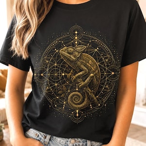 Puede incluir: Una camiseta negra de manga corta con un gráfico dorado de un camaleón centrado dentro de un complejo patrón geométrico de círculos y líneas entrelazadas. El diseño incluye el texto Low Energy Animals en la esquina inferior izquierda.