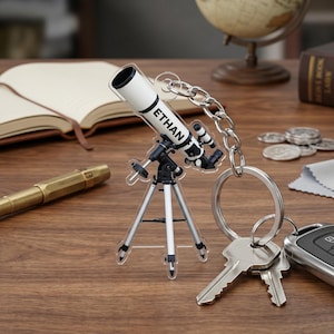 May include: A clear acrylic keychain featuring a miniature telescope design. The telescope is white and black, mounted on a small tripod. The name "ETHAN" is printed on the telescope. The keychain is attached to a chain and key ring, with keys and a car remote.