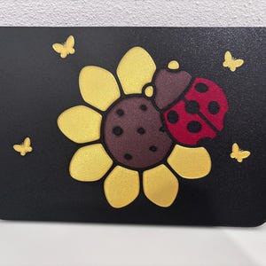 May include: A rectangular black plaque featuring a central yellow sunflower with a dark brown center. A red ladybug with black spots is positioned on the sunflower. Four small yellow butterflies are arranged around the flower design.