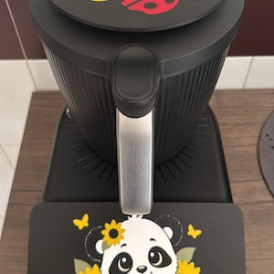 May include: A black kitchen appliance featuring a circular top lid decorated with a yellow sunflower and a red ladybug. A rectangular base mat sits in front, displaying a white and black panda bear surrounded by yellow sunflowers and butterflies.