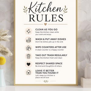 May include: A framed sign with the words "Kitchen RULES" in elegant script. Below, a list of kitchen rules is displayed, including "Clean as you go" and "Wash & put away dishes". The sign has a gold frame and is set against a neutral background.