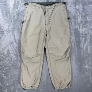 Pode incluir: Calças bege com fecho de correr frontal, bolsos laterais e cordões ajustáveis na cintura e nas bainhas. As calças apresentam costuras escuras visíveis ao longo das junções, proporcionando um visual estruturado com um corte descontraído.