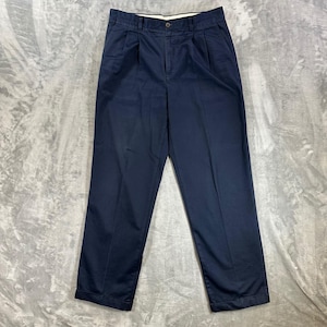 May include: A pair of navy blue trousers featuring a pleated front design, a button and zipper fly closure, and belt loops at the waistband. These full-length pants have a straight leg cut and a clean, solid-colored finish throughout the garment.