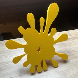 May include: A bright yellow, splash-shaped wall decoration. The design features multiple rounded extensions, resembling paint splatters. A keyhole is visible on the back for hanging. The object is made of a solid material and is set against a light wood surface.