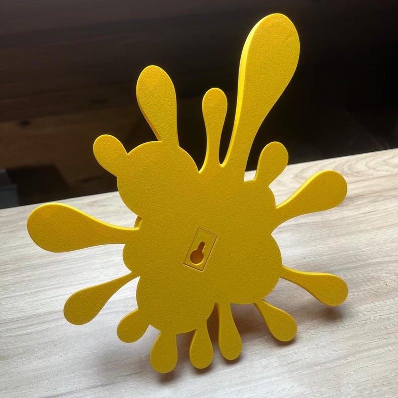 May include: A bright yellow, splash-shaped wall decoration. The design features multiple rounded extensions, resembling paint splatters. A keyhole is visible on the back for hanging. The object is made of a solid material and is set against a light wood surface.