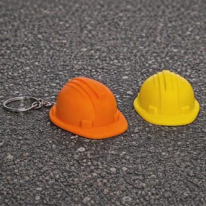 Può includere: Due caschi da cantiere in miniatura, uno arancione e uno giallo, sono mostrati. Il casco arancione è attaccato a un portachiavi in metallo argentato, mentre il casco giallo è separato. Entrambi gli oggetti presentano un design classico a coste.