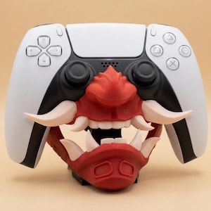 May include: A white and black PlayStation 5 controller with a red and white Oni mask design attached. The mask features sharp teeth and horns, adding a unique aesthetic to the gaming accessory.