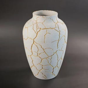 May include: A light gray ceramic vase with a speckled texture and a wide, rounded body. The exterior surface is covered in a network of irregular, branching gold-colored cracks that extend from the neck down to the base of the vessel.