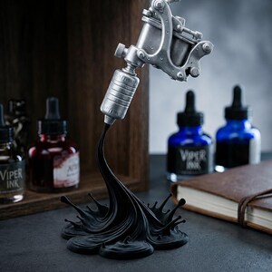 May include: A silver-colored tattoo machine sculpture with a black ink splash base. The sculpture is set against a backdrop of ink bottles and a wooden shelf. The bottles contain red and blue ink.