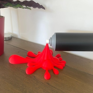 May include: A black spray paint can with a gray nozzle is mounted on a white wall, appearing to spray a red paint splatter onto a wooden surface. A red paint splatter is on the wooden surface.