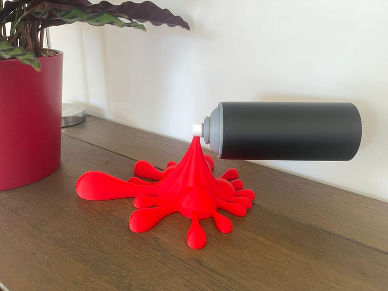 May include: A black spray paint can with a gray nozzle is mounted on a white wall, appearing to spray a red paint splatter onto a wooden surface. A red paint splatter is on the wooden surface.