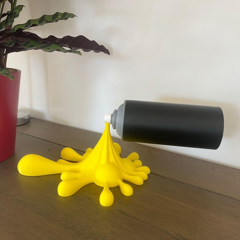 May include: A black spray paint can with a gray nozzle is attached to a yellow paint splatter. The can is mounted on a wall, and the yellow splatter sits on a wooden surface. A potted plant is in the background.