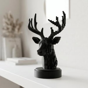Peut inclure: Une sculpture décorative noire en forme de tête de cerf. La tête de cerf est stylisée avec un design pixélisé sur le cou et la tête, et des bois lisses. La sculpture est montée sur une base ronde. La sculpture est exposée sur une surface blanche.