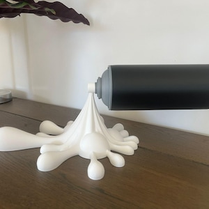 May include: A unique, wall-mounted toilet paper holder. The holder features a black cylindrical design, resembling a spray paint can, mounted on a white, stylized splash. The holder is designed to be mounted on a wall, and is made of a durable material.