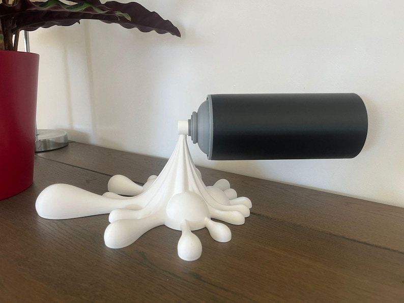 May include: A unique, wall-mounted toilet paper holder. The holder features a black cylindrical design, resembling a spray paint can, mounted on a white, stylized splash. The holder is designed to be mounted on a wall, and is made of a durable material.