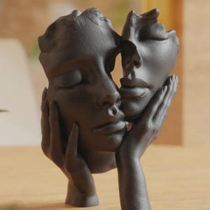 Peut inclure: Une sculpture marron foncé représentant deux visages dans une étreinte tendre. Les visages ont les yeux fermés et des traits délicats, tenus par une paire de mains. C'est une pièce décorative.