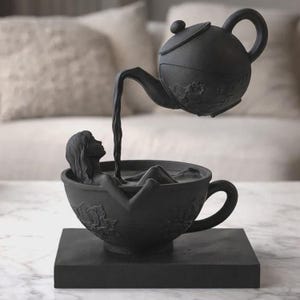 Peut inclure: Sculpture noire représentant une femme dans une tasse à thé, avec une théière versant du liquide sur elle. La tasse et la théière sont ornées de motifs floraux. L'ensemble repose sur une base carrée.