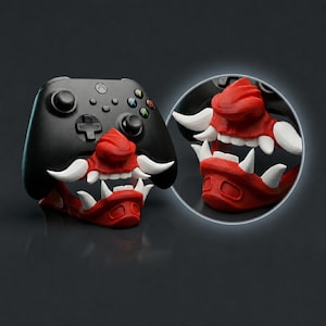 May include: Black Xbox controller with a red and white Oni mask design attached. The mask features sharp white fangs and a detailed red face. A circular inset shows a close-up of the mask. Gaming accessory.