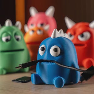 Peut inclure: Plusieurs organisateurs de câbles en forme de monstres colorés. Le monstre bleu tient un câble noir dans sa bouche. Les autres monstres sont verts, orange, roses et rouges. Un petit appareil noir est au premier plan.
