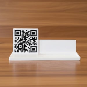 May include: A white rectangular display stand featuring a square black and white QR code on the left side. The stand has a flat base and a vertical back panel, providing a clean, minimalist structure for signage or information display purposes.