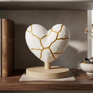 May include: A white heart-shaped sculpture featuring gold-coloured cracks in the kintsugi style, mounted on a cylindrical post and a round, light-coloured wooden base. The object is positioned next to a brown book with the word KINTSUGI on the spine.