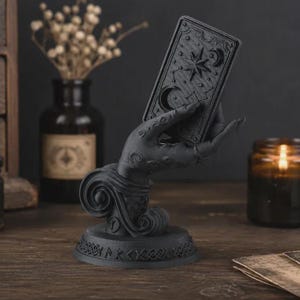May include: A matte black sculpture of a hand emerging from a swirling base, holding a rectangular tarot card featuring embossed celestial motifs including a star, crescent moon, and constellations. The circular base is decorated with runic symbols.