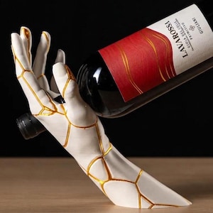 May include: A white hand-shaped wine bottle holder featuring gold-colored cracks across the surface. The holder supports a bottle of Lavarossa Gioia del Colle Primitivo wine with a red and white label. The design mimics a kintsugi aesthetic.
