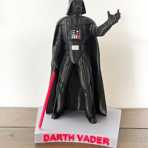 May include: A black Darth Vader figurine with a red lightsaber, on a white base. The base reads "Darth Vader" in red. The figurine is a detailed representation of the Star Wars character, a popular film franchise.
