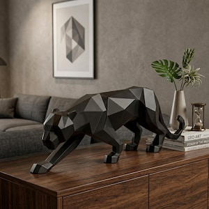 May include: A black panther sculpture featuring a geometric, faceted design. The panther is depicted in a low, prowling stance with its head lowered and tail curved upward. The surface is smooth and matte, reflecting light on its sharp edges.