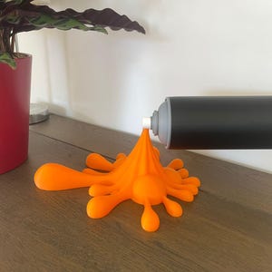 May include: A black and gray spray paint can with an orange paint splatter design. The can is mounted on a white wall, and the orange paint appears to be flowing onto a wooden surface. A red plant pot is in the background.