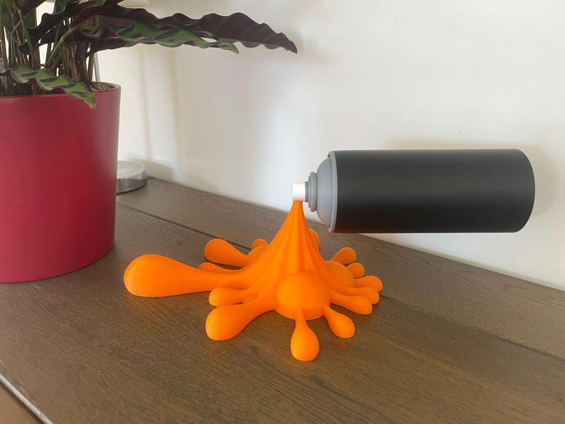 May include: A black and gray spray paint can with an orange paint splatter design. The can is mounted on a white wall, and the orange paint appears to be flowing onto a wooden surface. A red plant pot is in the background.