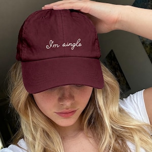 May include: A maroon baseball cap featuring the white embroidered text "I'm single" in a cursive script font across the front panel. The cap has a structured crown, a curved brim with visible stitching, and a button at the top center of the crown.