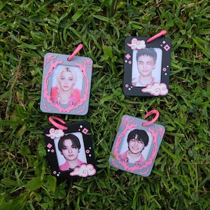 May include: Four square-shaped photo charms with pink accents and images of people. Each charm has a pink loop at the top. The frames are gray and black with cloud and decorative accents. The charms are on a bed of green grass.
