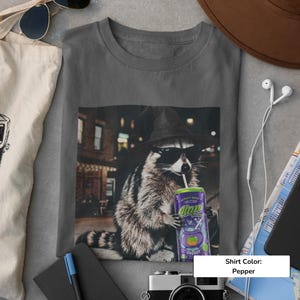 May include: A pepper-colored t-shirt featuring a graphic print of a raccoon wearing a witch hat and sunglasses while drinking from a purple can labeled Alani Nu. The raccoon is depicted in an urban setting at night with blurred city lights.
